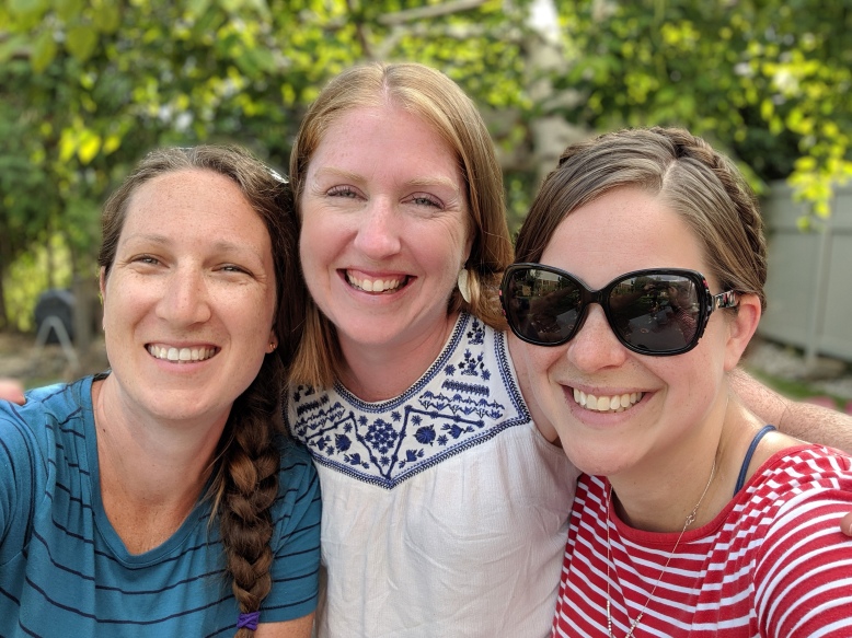 Three women, close together, smile at the camera. It’s sunny outside and there are trees in the background. 