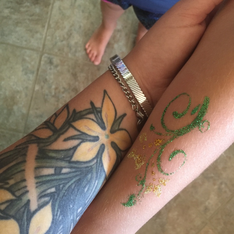 Close up of two arms, an adult’s arm with a real tattoo of green and yellow flowers, and a child’s arm with a temporary glitter tattoo of green and yellow flowers. 