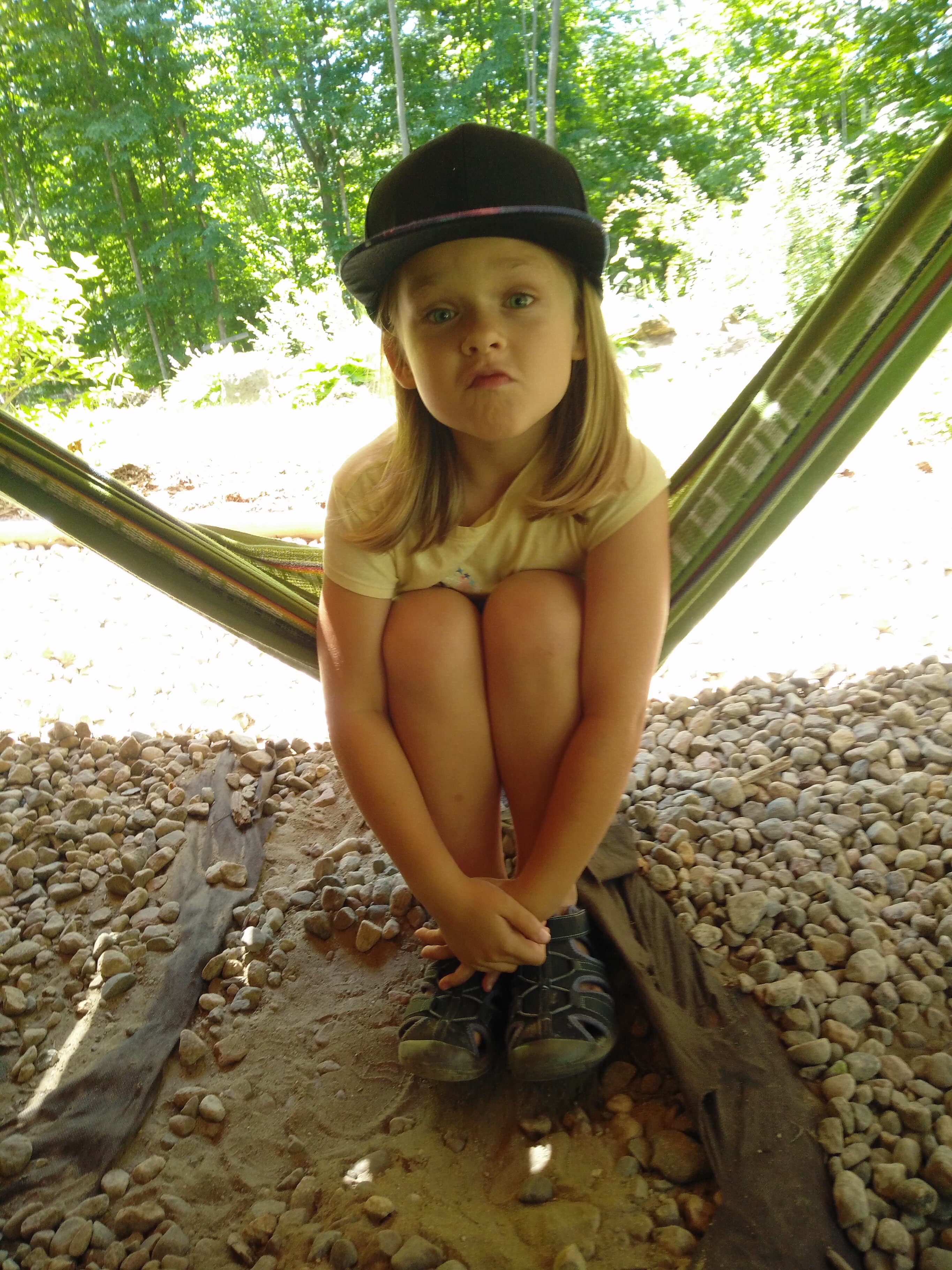 A 6 year old girl sits in a hammock and makes a silly face at the camera. 