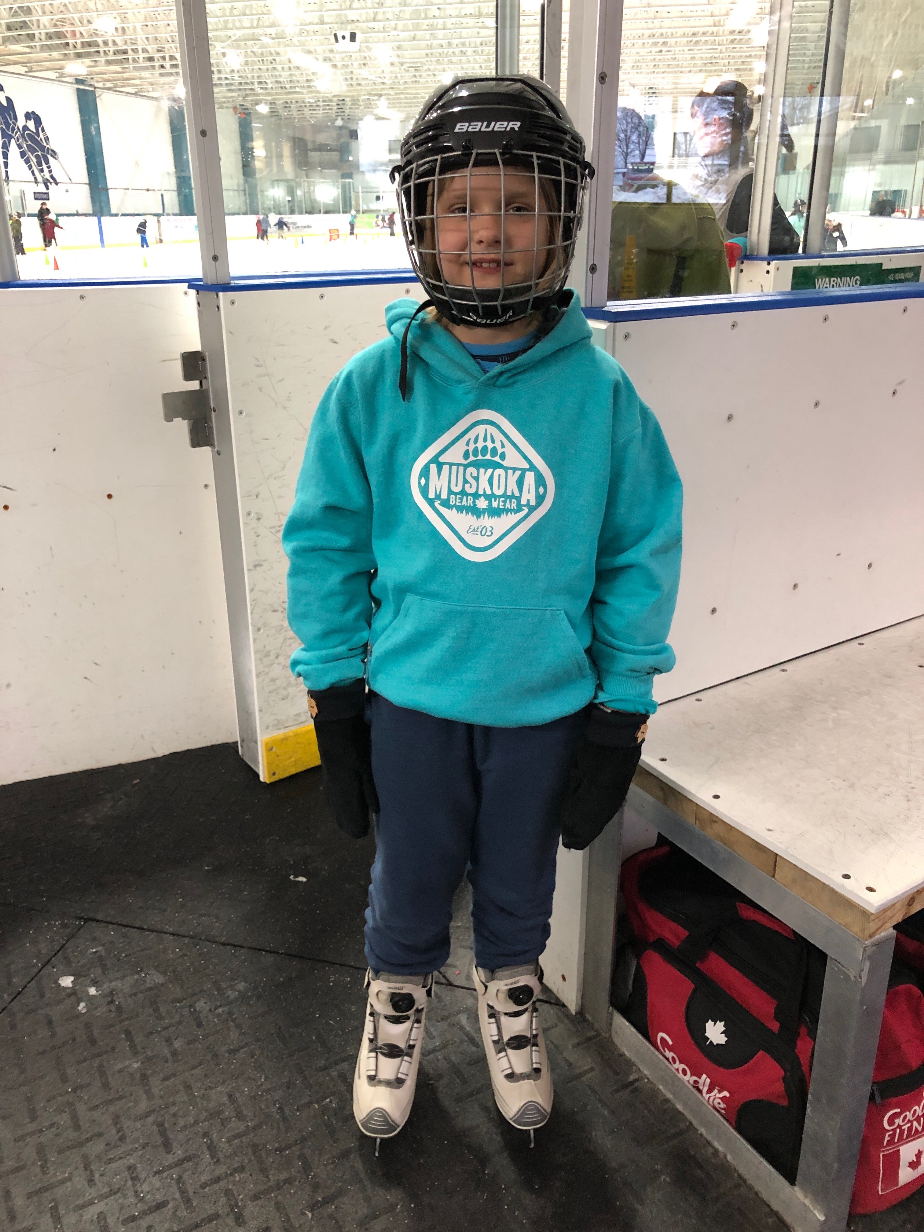 An 8 year old blonde girl stands in front of the ice in a skating rink. She is wearing skates, an aqua sweatshirt, and a hockey helmet with a wire face mask. 