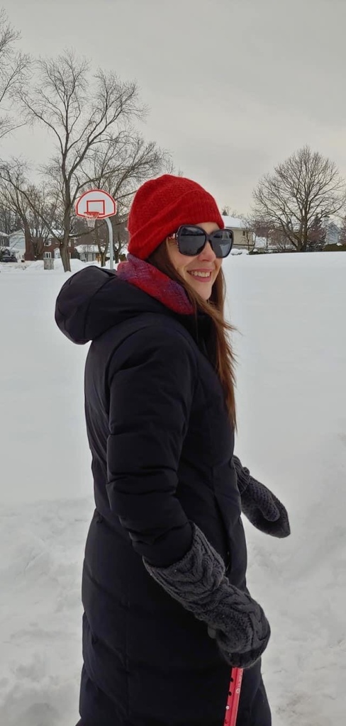 Jasmine is walking in a snowy field wearing a black coat, black sunglassses,  and a red hat.  She is carrying a red cane and smiling at the camera. 